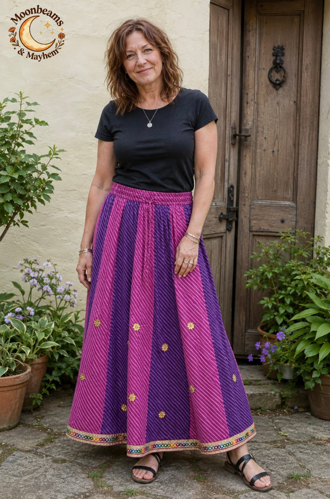A woman wearing a long pleated skirt with vertical stripes of magenta pink and purple, decorated with small gold floral motifs and a colorful border hem, standing in front of a rustic wooden door.